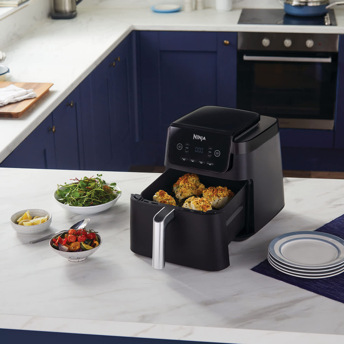 Airfryer avec tiroir ouvert pos&eacute; sur une table, contenant du poisson, entour&eacute; de plusieurs assiettes de l&eacute;gumes.