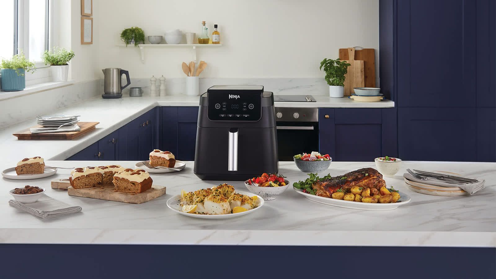 Airfryer pos&eacute; sur une table, entour&eacute; de plusieurs assiettes de nourriture, notamment du poisson, un g&acirc;teau, ainsi que de la viande et des pommes de terre.