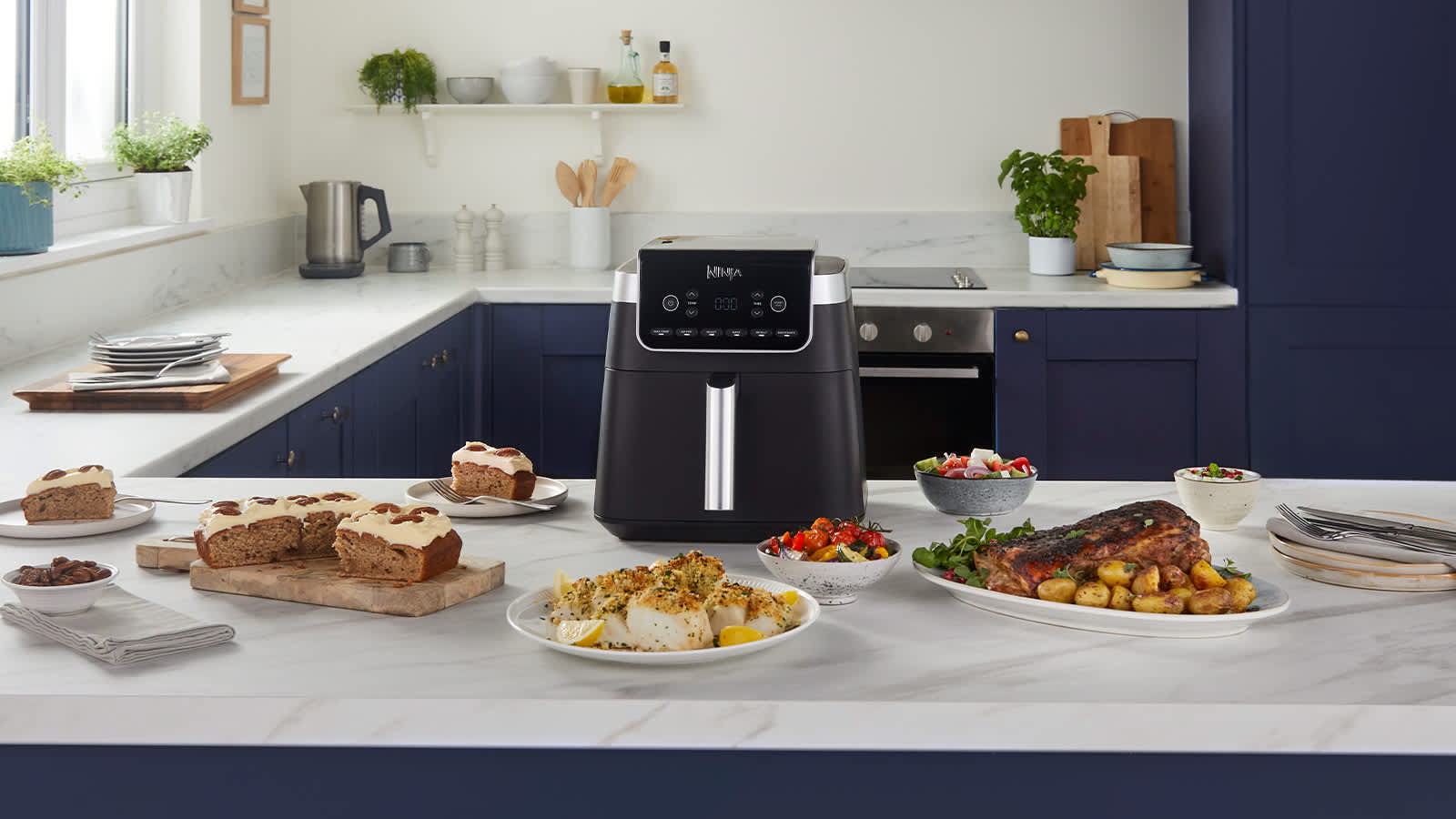 Airfryer pos&eacute; sur une table, entour&eacute; de plusieurs assiettes de nourriture, notamment du poisson et un g&acirc;teau.