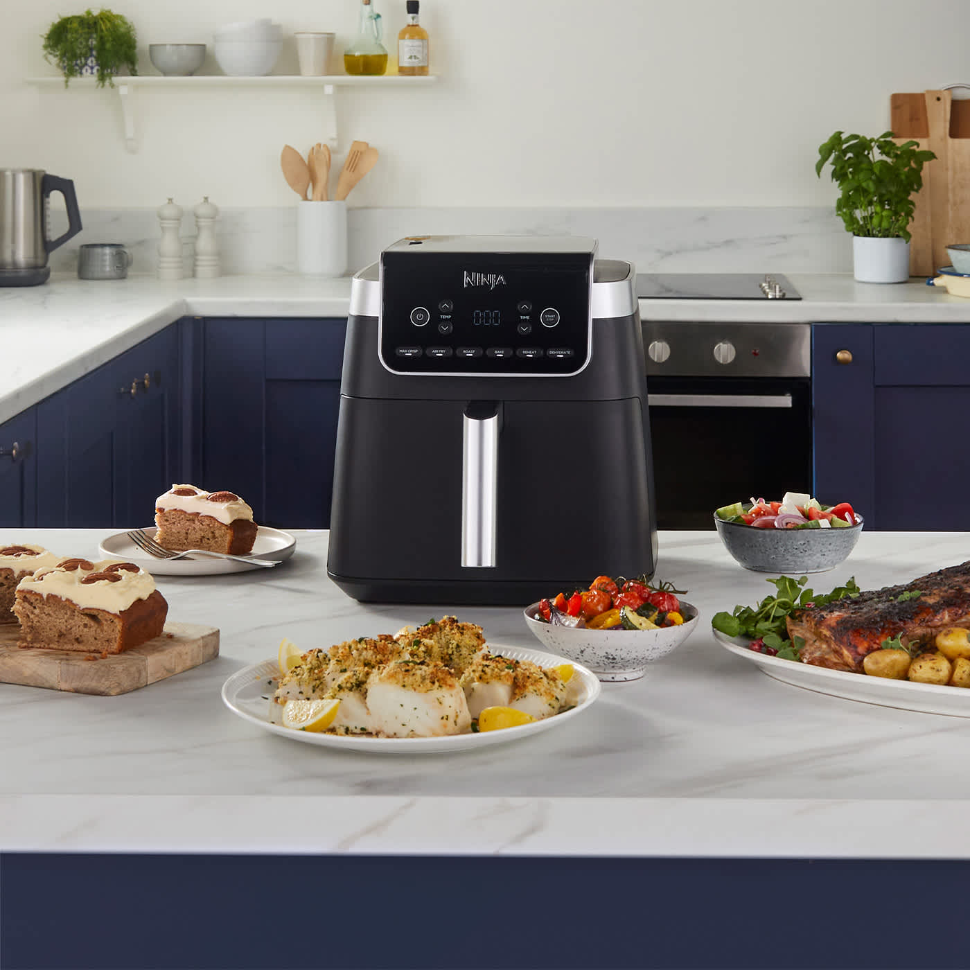 Airfryer pos&eacute; sur une table, entour&eacute; de plusieurs assiettes de nourriture, notamment du poisson et un g&acirc;teau.