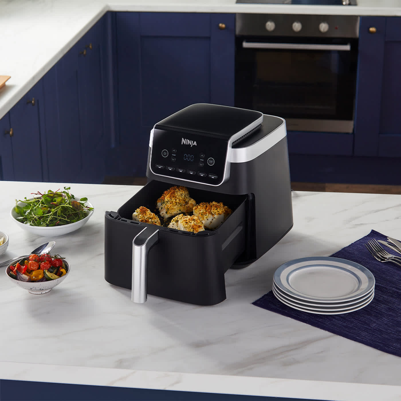 Airfryer avec tiroir ouvert contenant du poisson, entour&eacute; de plusieurs assiettes de l&eacute;gumes.