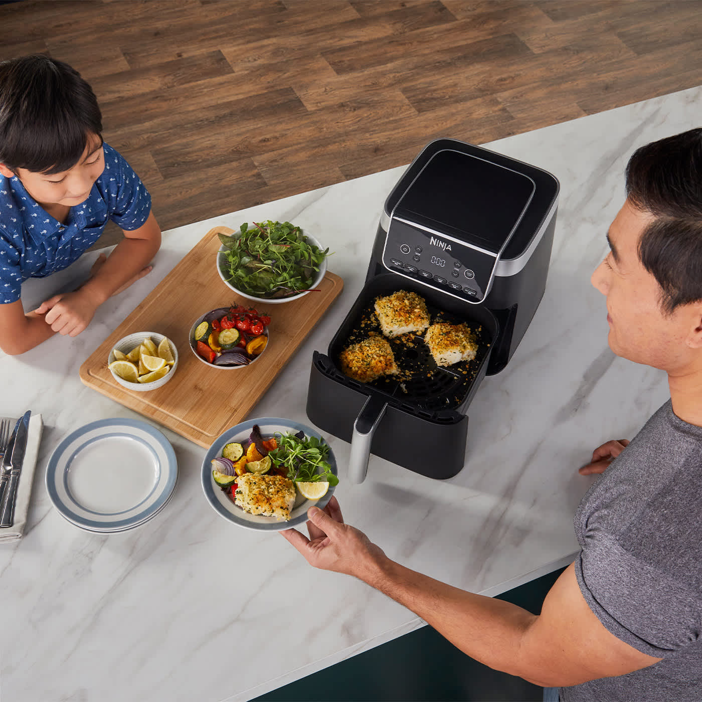 Airfryer avec tiroir ouvert contenant du poisson, un p&egrave;re servant une assiette &agrave; son fils, avec des plats de l&eacute;gumes sur la table.