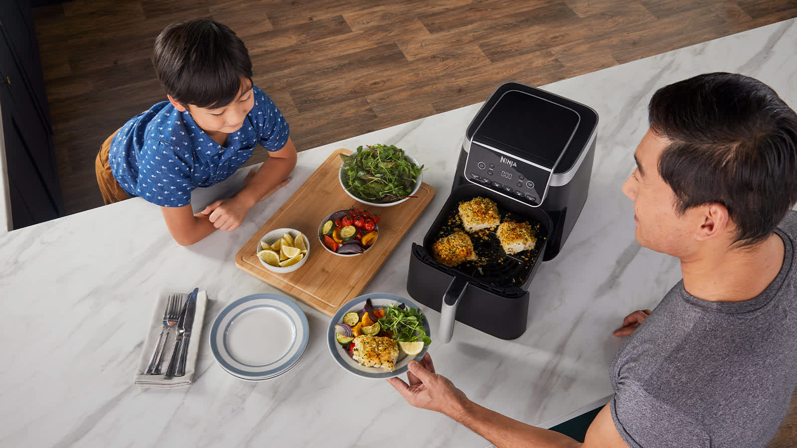 Airfryer avec tiroir ouvert contenant du poisson, un p&egrave;re servant une assiette &agrave; son fils, avec des plats de l&eacute;gumes sur la table.