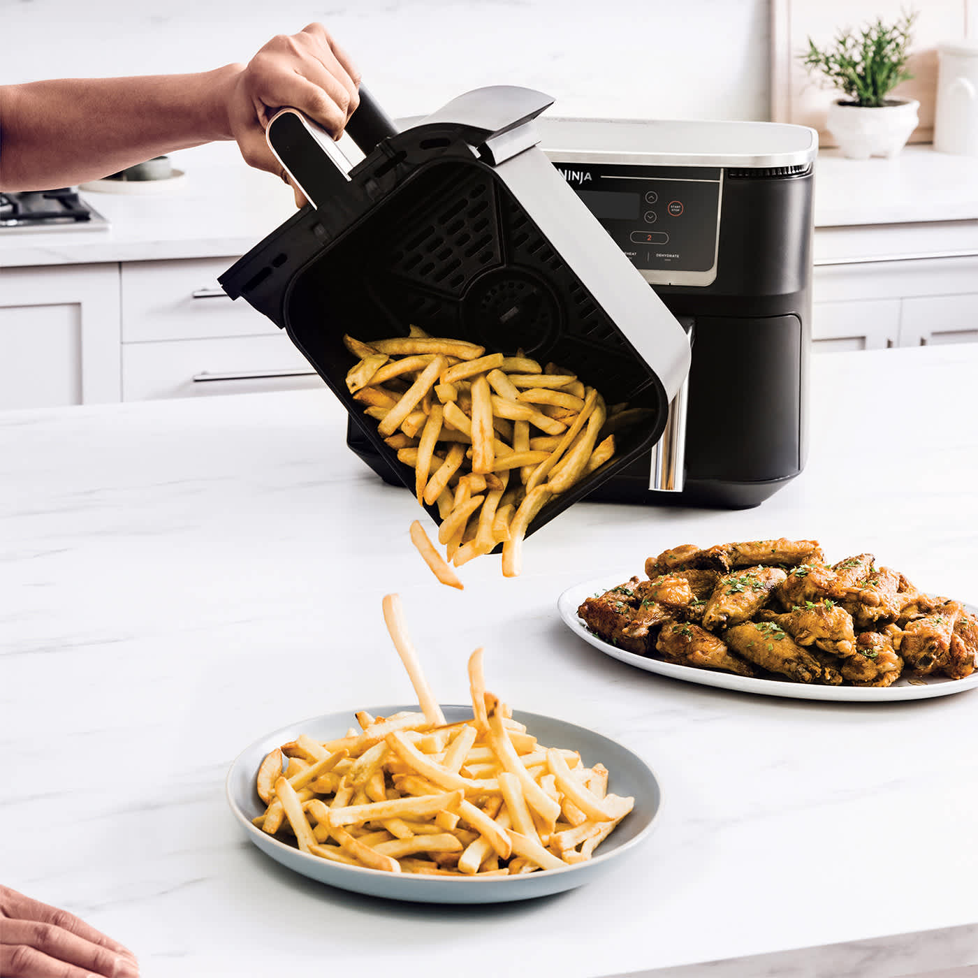 Personne versant des frites d'un tiroir d'air fryer dans une assiette, avec une assiette d'ailes de poulet en arri&egrave;re-plan.