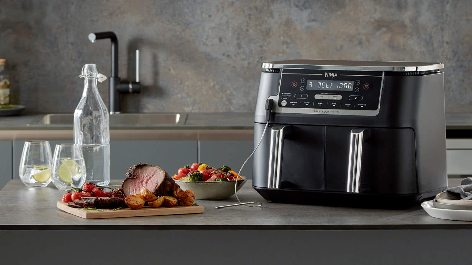 Air fryer Ninja avec thermosonde sur un comptoir de cuisine, accompagn&eacute; d'un r&ocirc;ti, d'un bol de l&eacute;gumes et d'une bouteille d'eau.