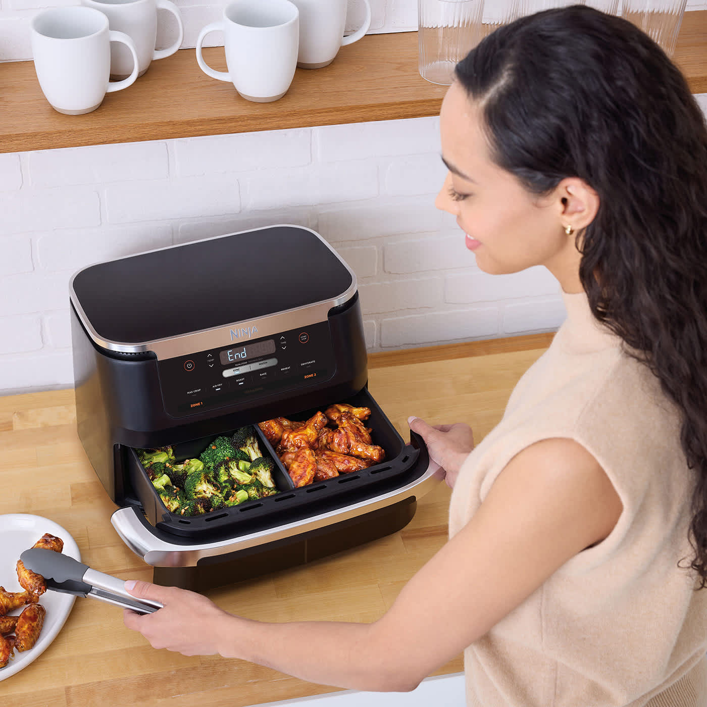 Femme ouvrant le tiroir de l'air fryer Ninja avec du brocoli d'un c&ocirc;t&eacute; et des ailes de poulet de l'autre.