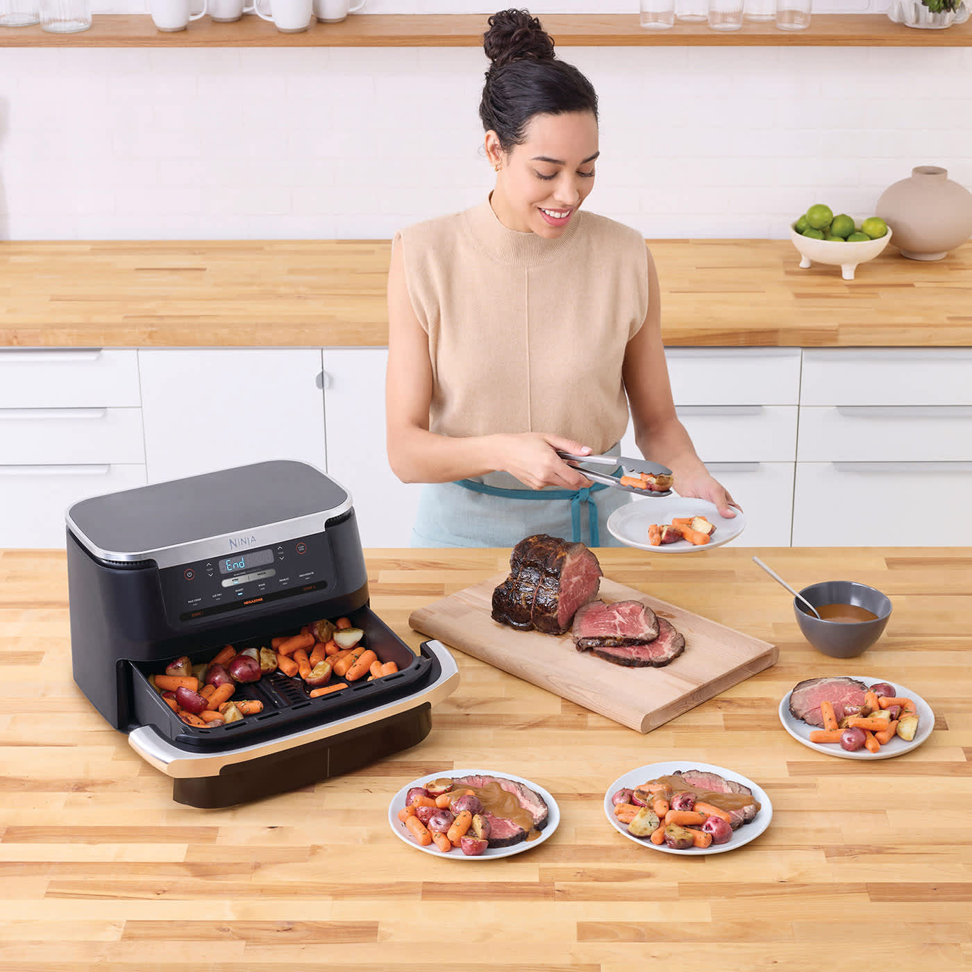 Air fryer contenant des l&eacute;gumes sur un comptoir de cuisine, femme servant des l&eacute;gumes dans une assiette, table avec des plats de viande et l&eacute;gumes et une planche &agrave; d&eacute;couper avec un r&ocirc;ti de b&oelig;uf.