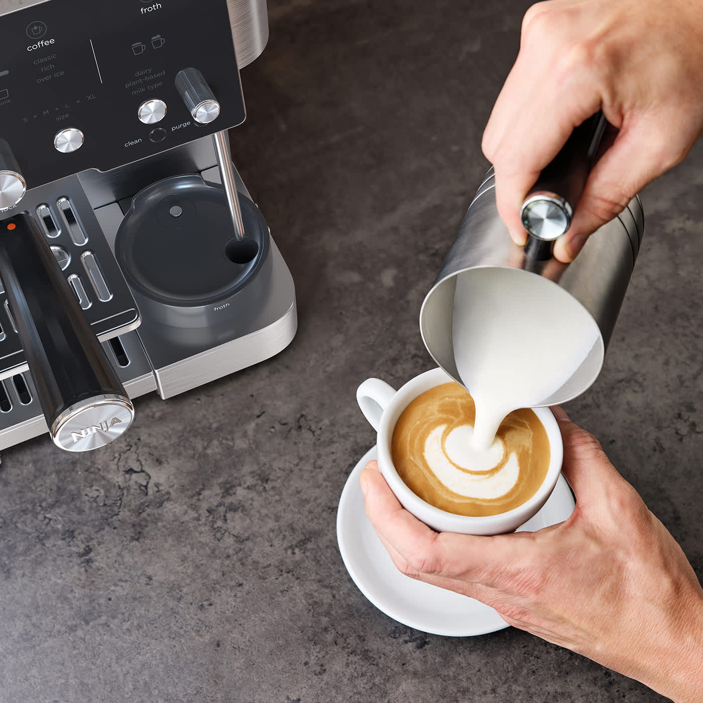 Personne versant de la mousse de lait dans un caf&eacute;, avec une machine &agrave; caf&eacute; en arri&egrave;re-plan.