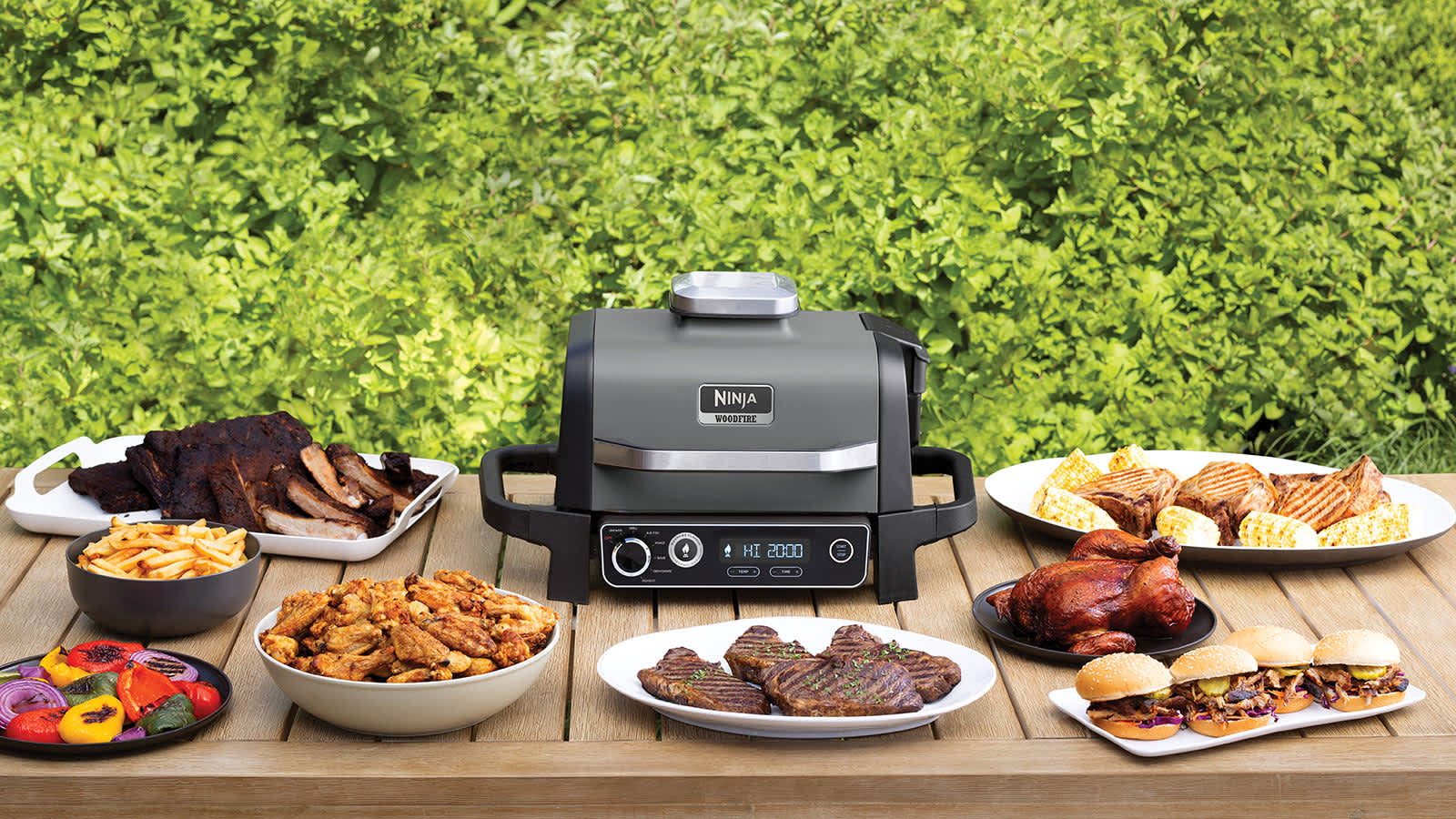 Barbecue Ninja OG701EU sur une table entour&eacute; de plats de steaks, c&ocirc;tes lev&eacute;es et poulet.
