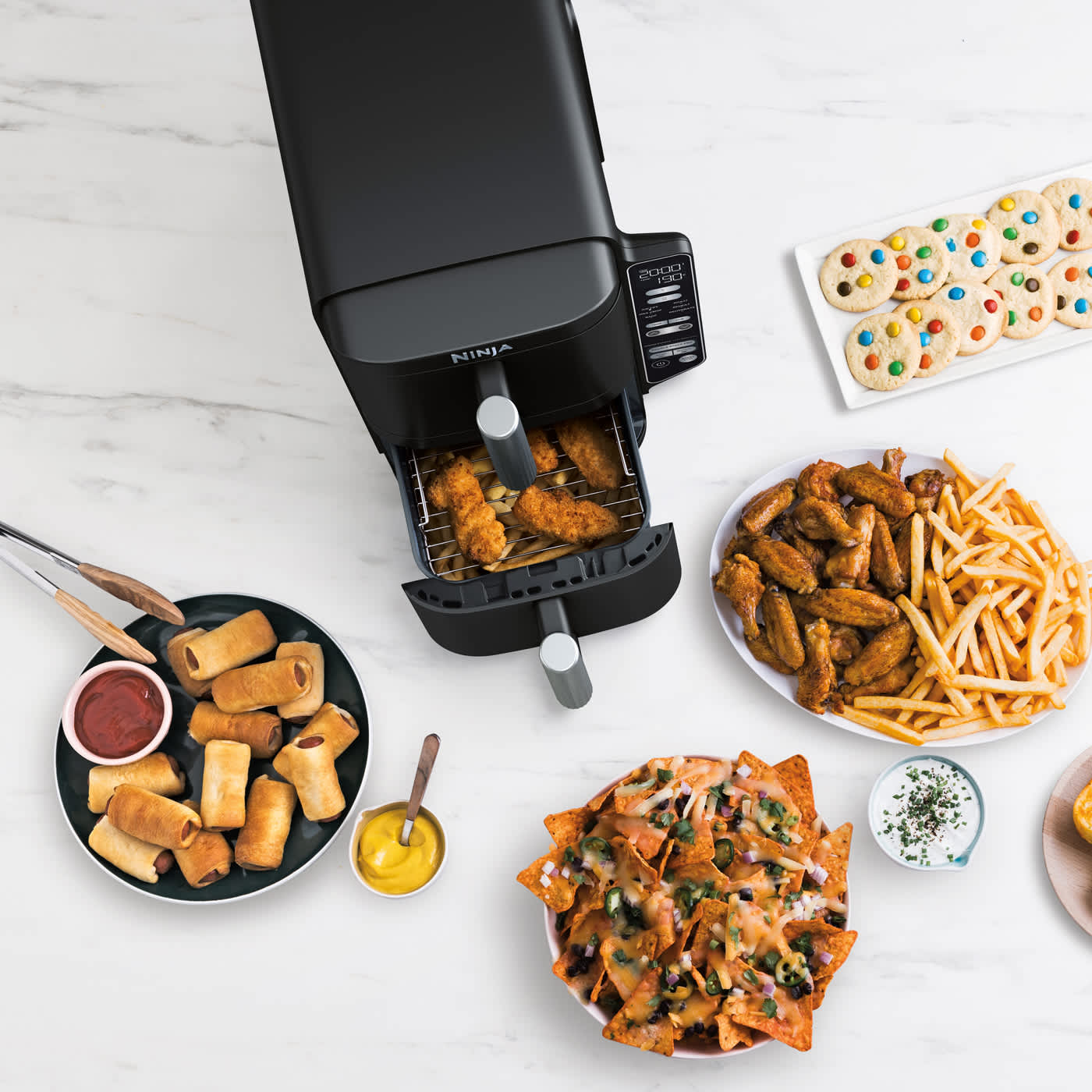 Airfryer avec tiroir ouvert contenant du poulet, pos&eacute; sur un plan de travail, entour&eacute; de plusieurs assiettes de nourriture, notamment des nachos, du poulet avec des frites et des cookies.