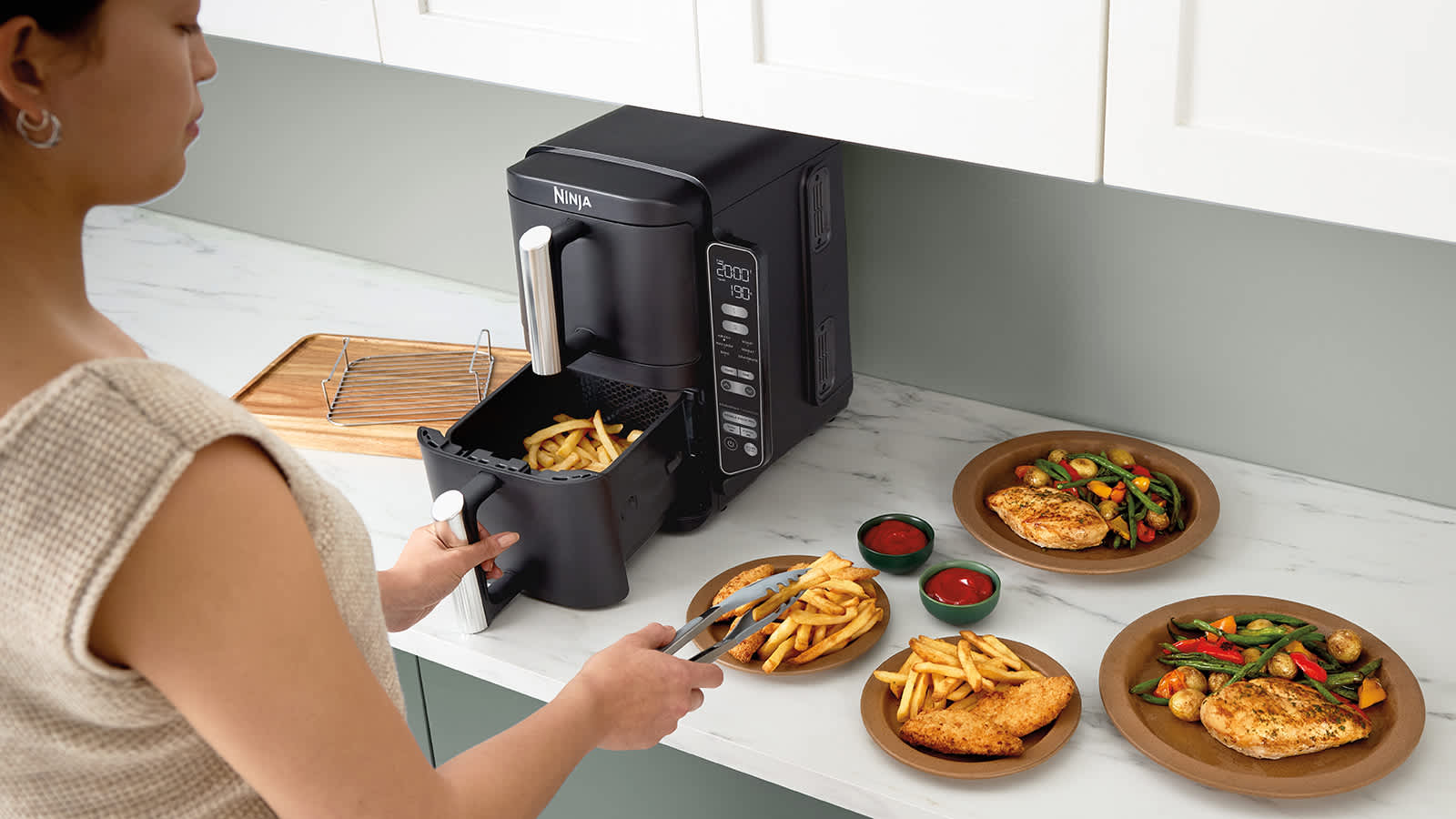 Personne retirant des frites de la friteuse &agrave; air, pos&eacute;e sur un plan de travail &agrave; c&ocirc;t&eacute; de plusieurs assiettes de nourriture.