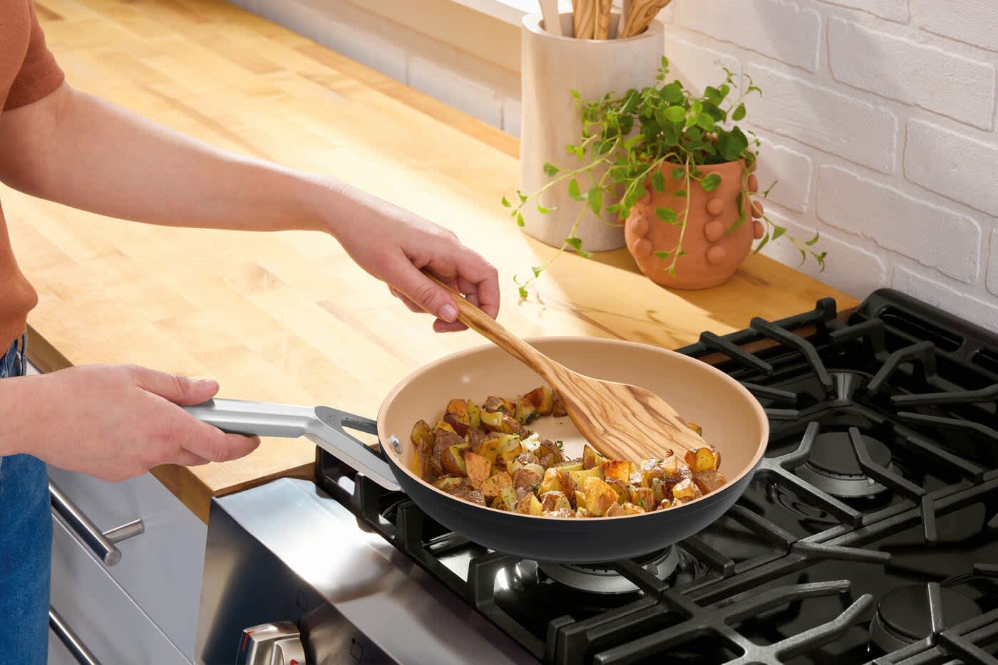Beschichtete Bratpfanne auf Gasherd mit angebratenen Kartoffeln und Kr&auml;utern, gewendet mit Holzl&ouml;ffel