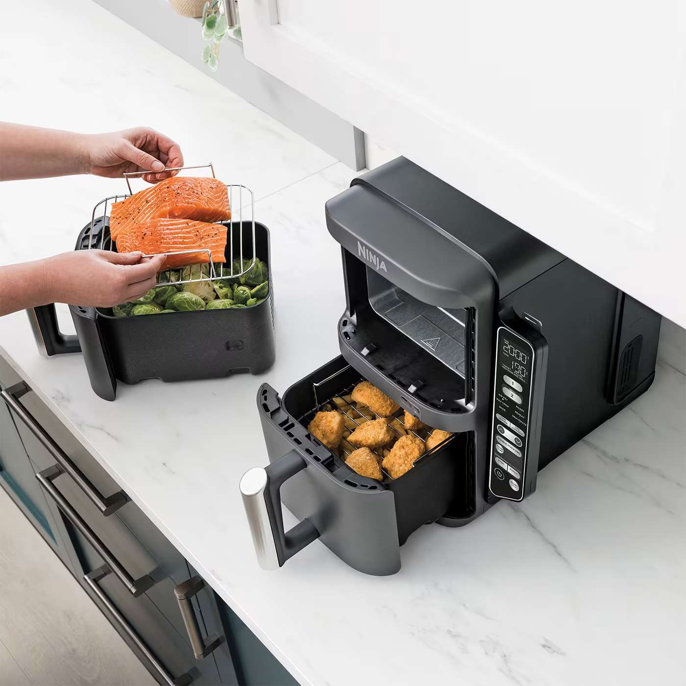 Personne cuisinant du saumon et des l&eacute;gumes dans un tiroir et du poulet dans l'autre tiroir de l'air fryer Ninja.