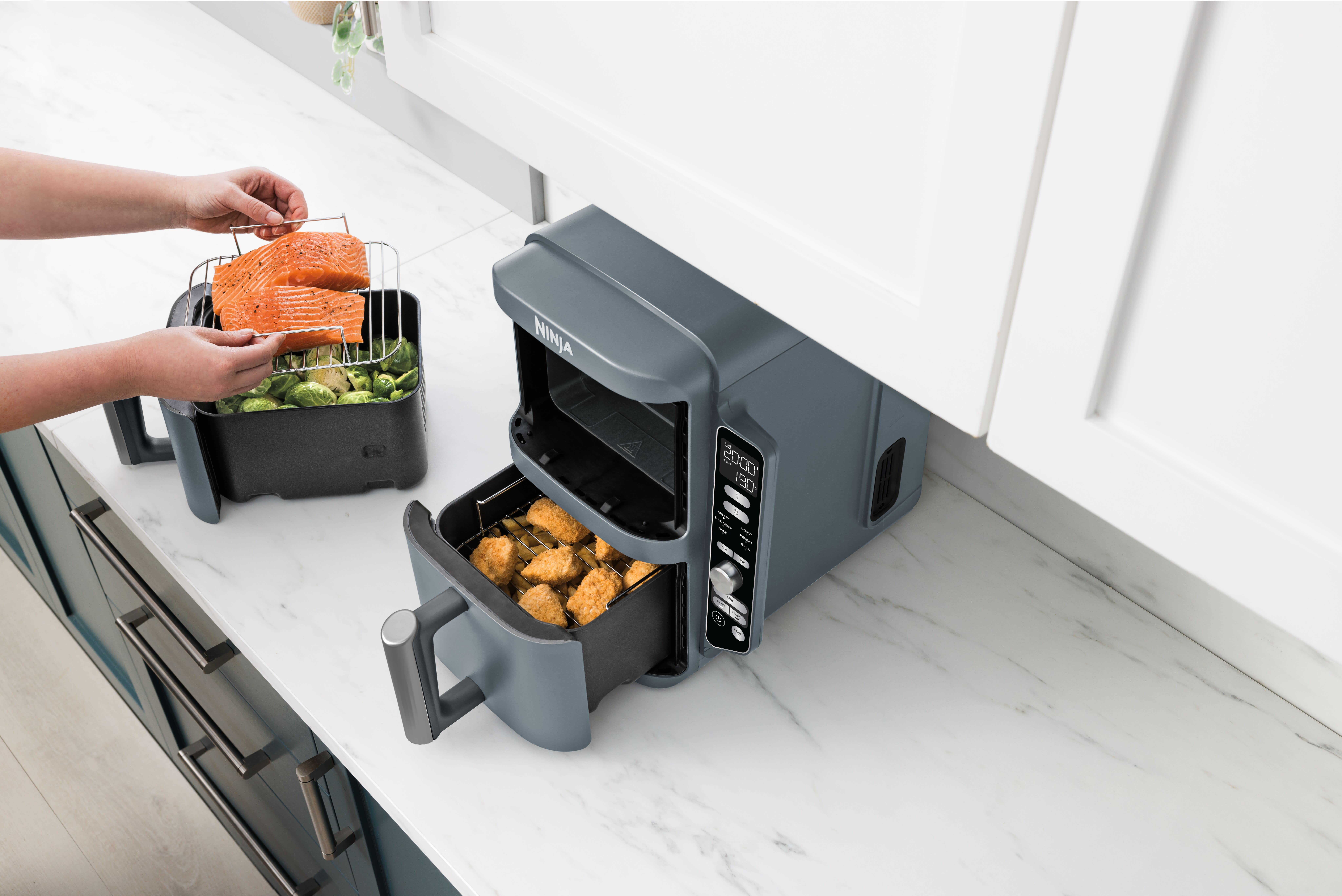 Personne disposant du saumon sur une grille empilable et par dessus des l&eacute;gumes dans un tiroir et du poulet dans l'autre tiroir de l'air fryer Ninja.