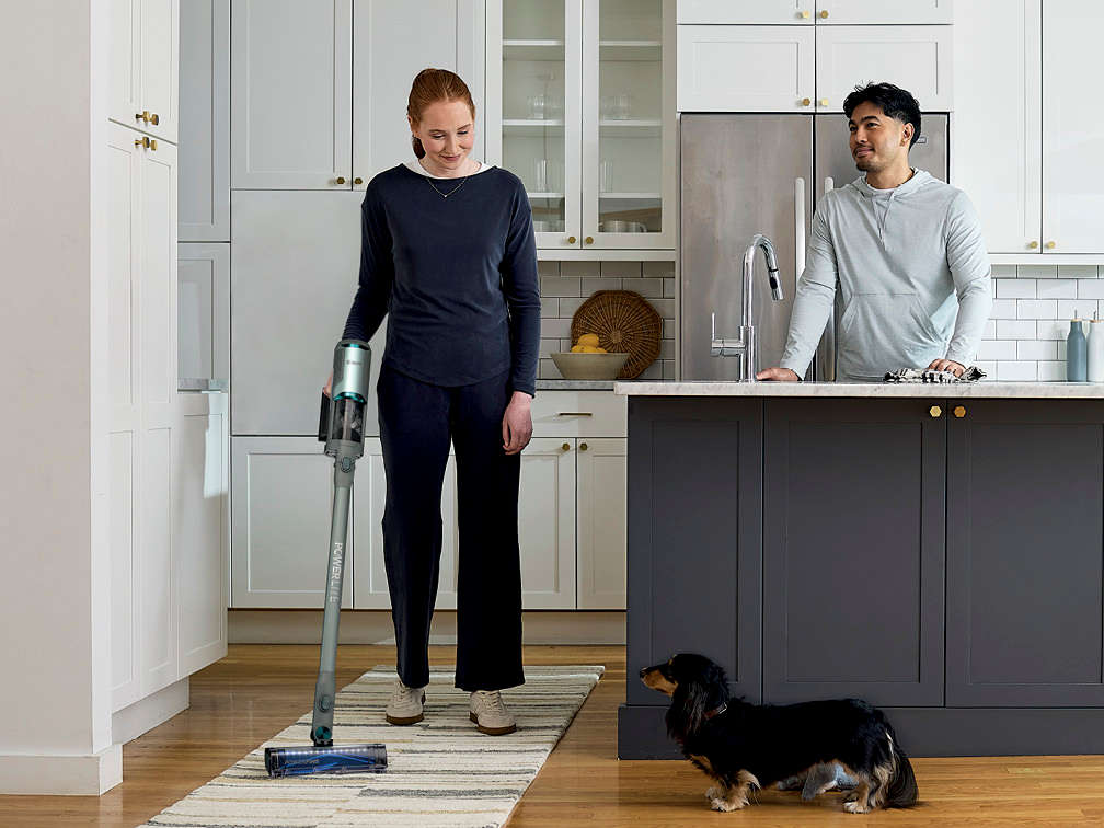 Kobieta odkurza dywanik odkurzaczem bezprzewodowym Shark PowerLite Clean & Empty Pet a obok przygląda się temu mężczyzna i pies