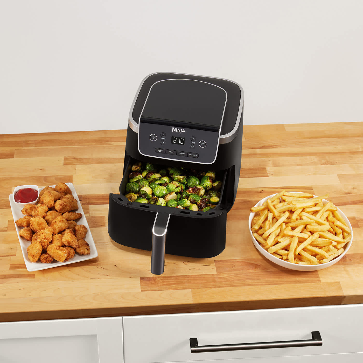 Immagine di una friggitrice ad aria Ninja 1 cassetto con dentro verdure e vicino un piatto con pollo e uno con patatine