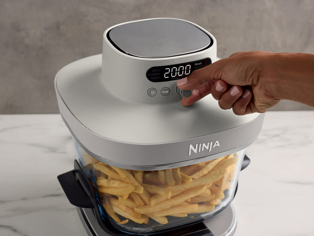 Osoba ustawia parametry pieczenia air fryera Ninja Crispi Pro AS101EU. W szklanym naczyniu CleanCrisp znajduje się duża porcja frytek