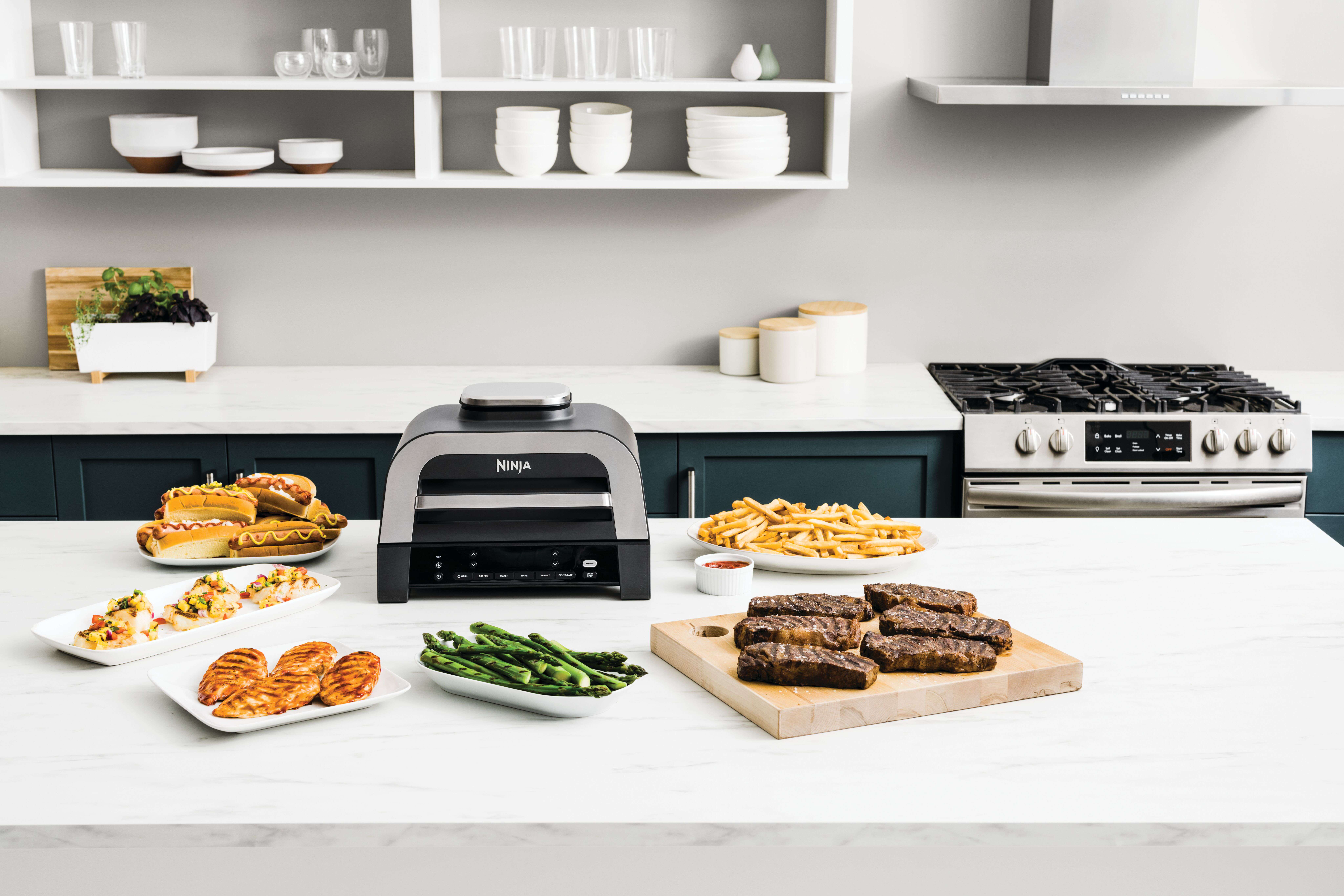 Grill i air fryer Ninja 6-w-1 Max DG551EU stojący na blacie kuchennym z wieloma potrawami obok
