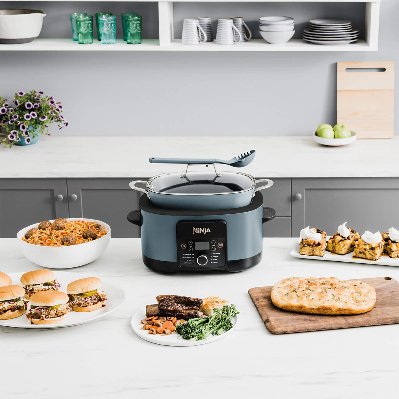 Wolnowar i multicooker Ninja Foodi MC1001EU stojący w otoczeniu wielu gotowych dań przygotowanych z jego udziałem
