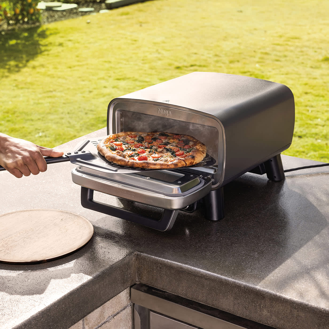 Osoba wyjmuje upieczoną pizzę z elektrycznego zewnętrznego pieca do pizzy i air fryera Ninja Artisan MO201EU