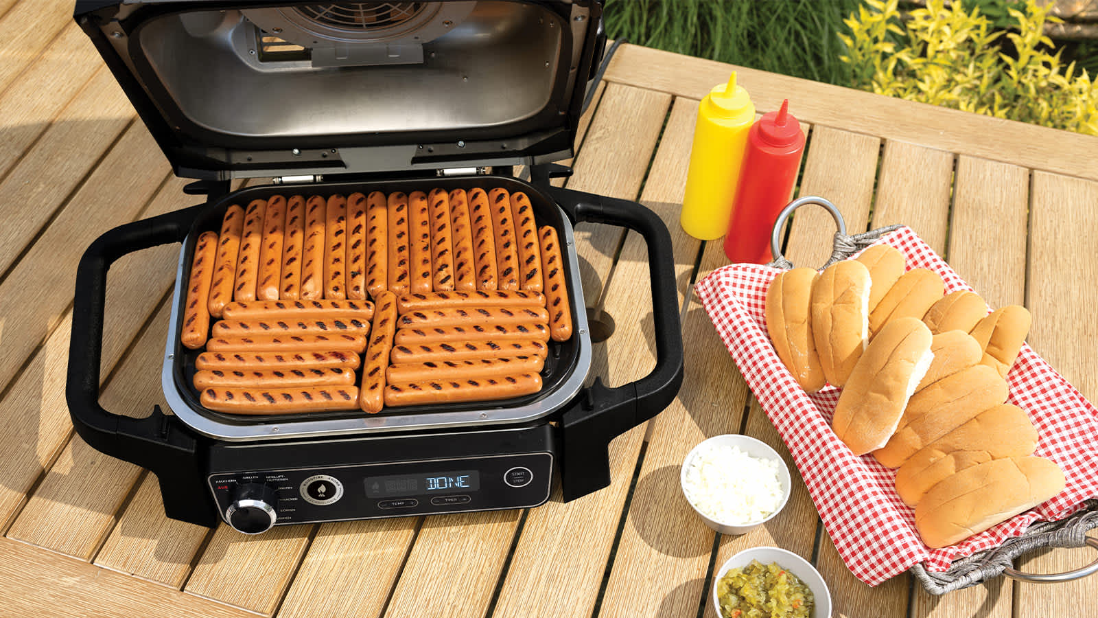Grill elektryczny Ninja OG701EU na stole z upieczonymi kiełbaskami