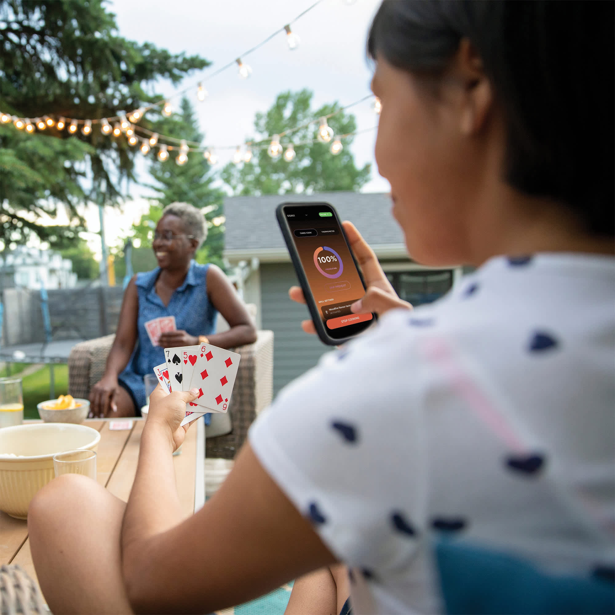 Wiele os&oacute;b gra ze sobą w karty, a jedna trzyma dodatkowo smartfon z ekranem aplikacji Ninja Pro Connect, aby na bieżąca podglądać parametry pracy grilla elektrycznegp Ninja Woodfire Pro Connect XL OG901EU