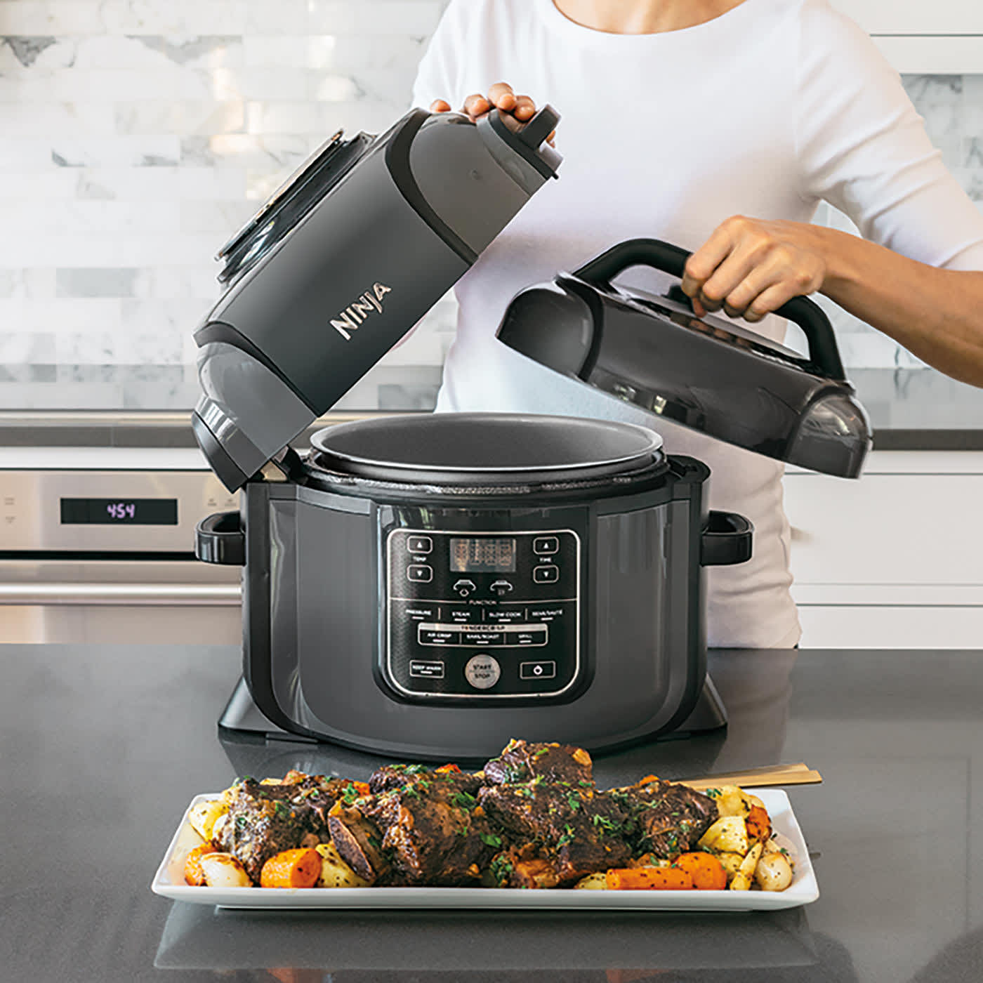 Kobieta przykrywa pokrywką szybkowar i multicooker Ninja. Przed nim stoi p&oacute;łmisek z upieczonym mięsem i dodatkami