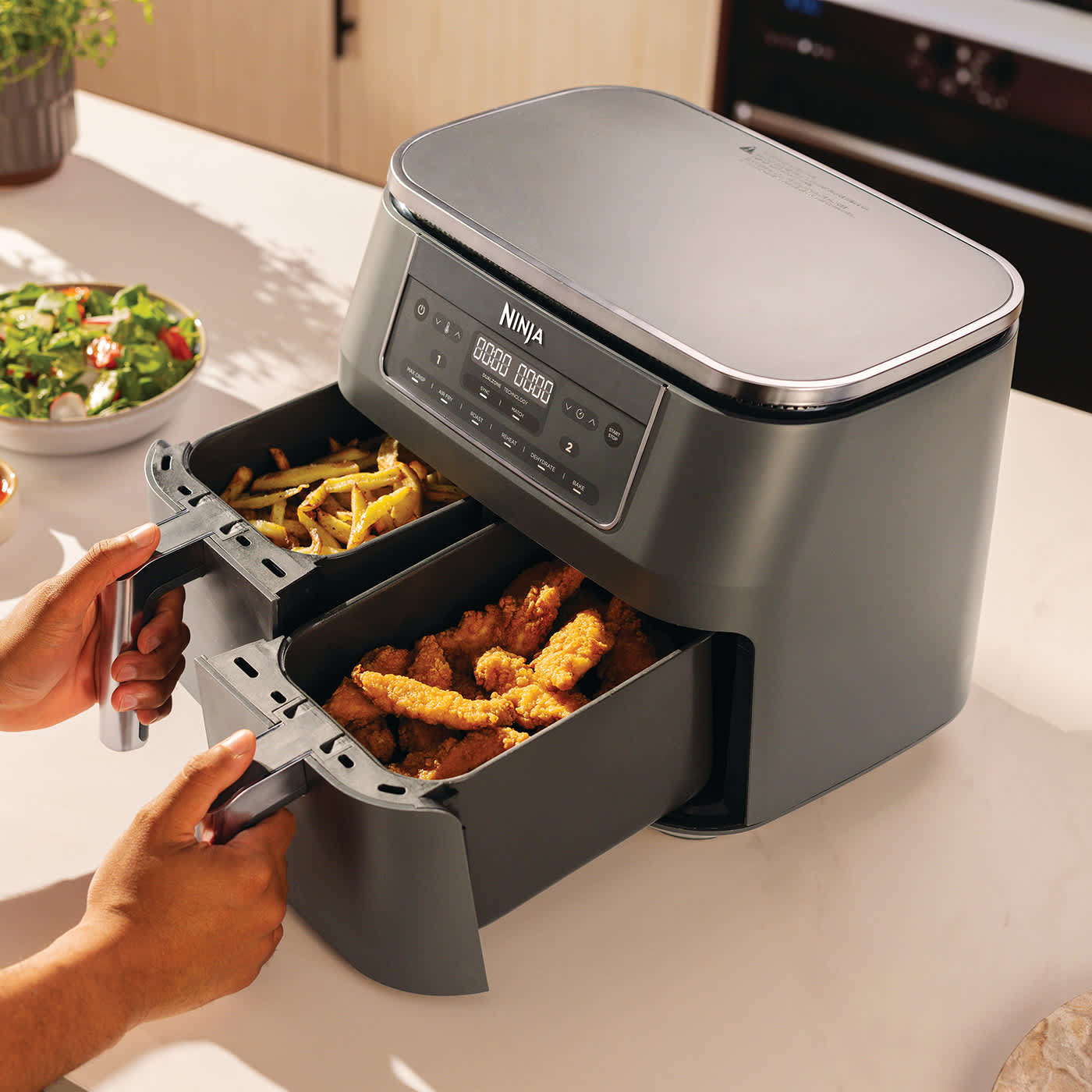Personne ouvrant les deux tiroirs de l'air fryer, l'un contenant du poulet et l'autre des frites.
