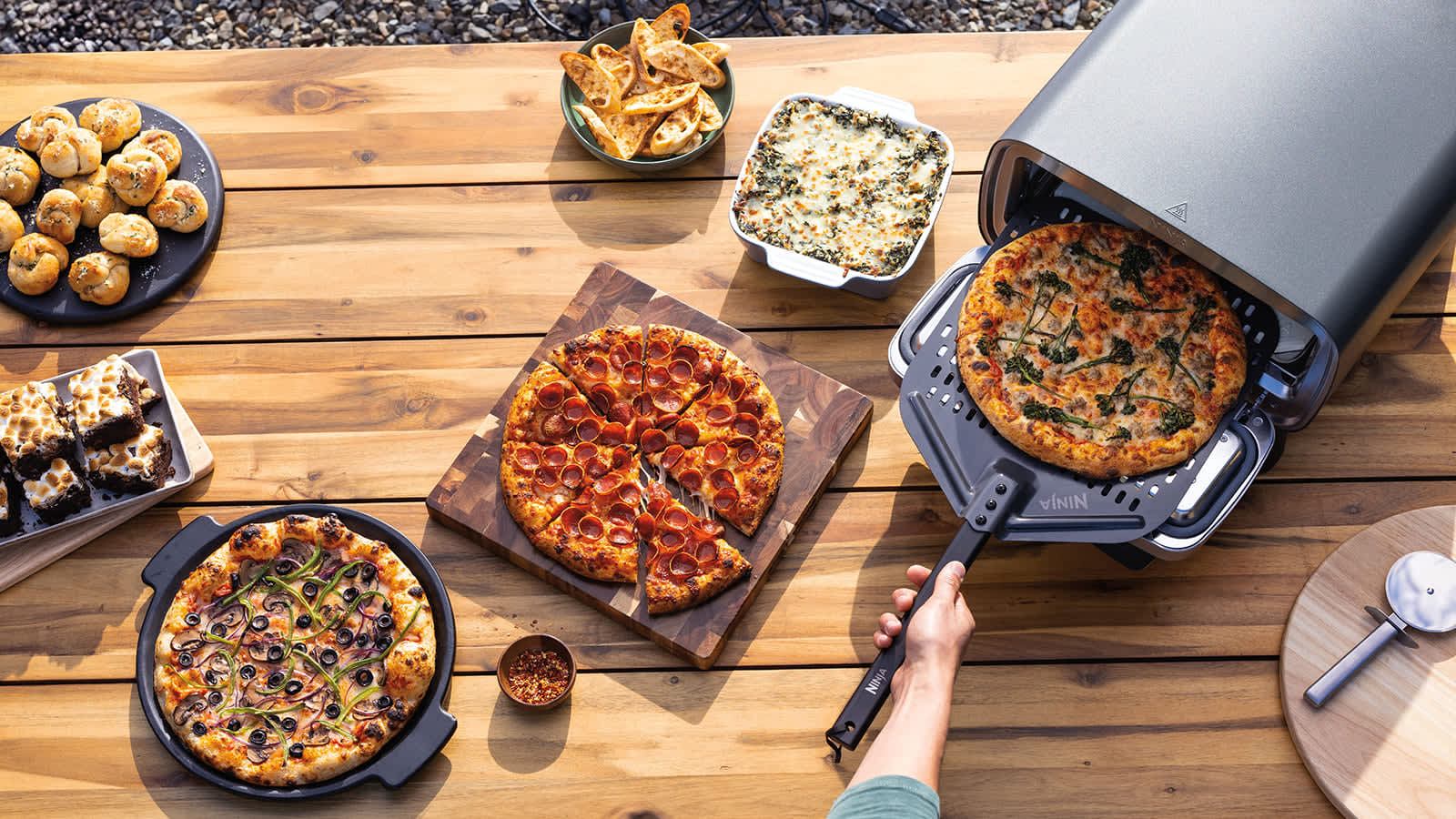 Personne mettant une pizza dans le four &agrave; pizza Ninja entour&eacute; d'assiettes d'ailes de poulet et d'une autre pizza.