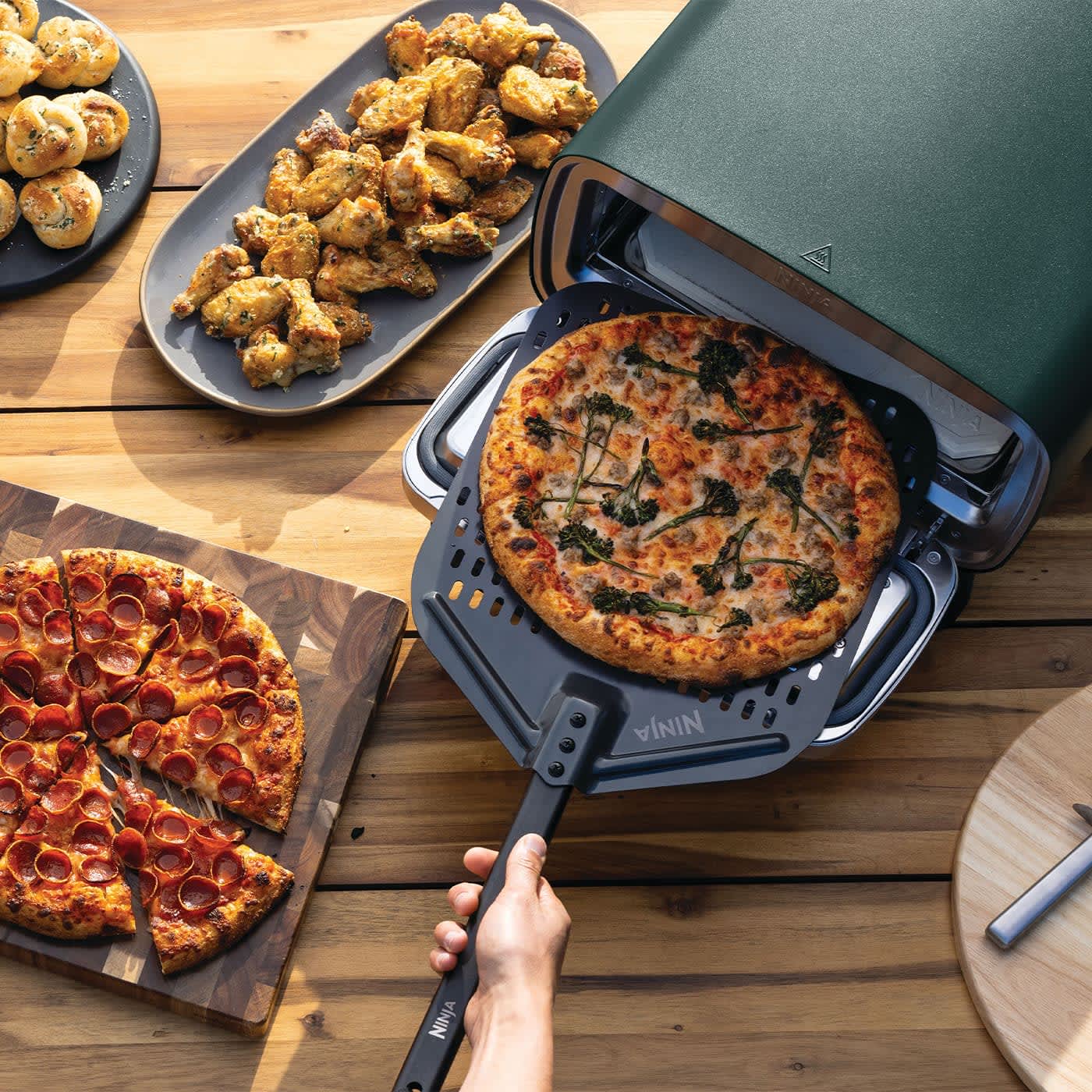 Personne mettant une pizza dans un four &agrave; pizza Ninja, entour&eacute; d&rsquo;autres pizzas et d&rsquo;ailes de poulet.