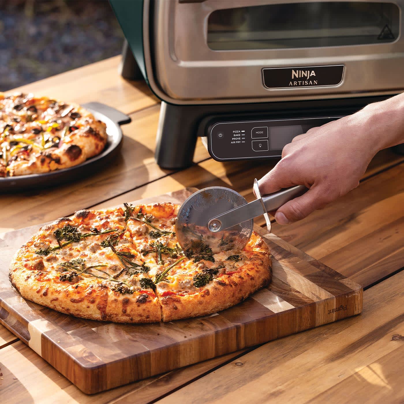 Personne coupant une pizza, avec un four &agrave; pizza Ninja en arri&egrave;re-plan.