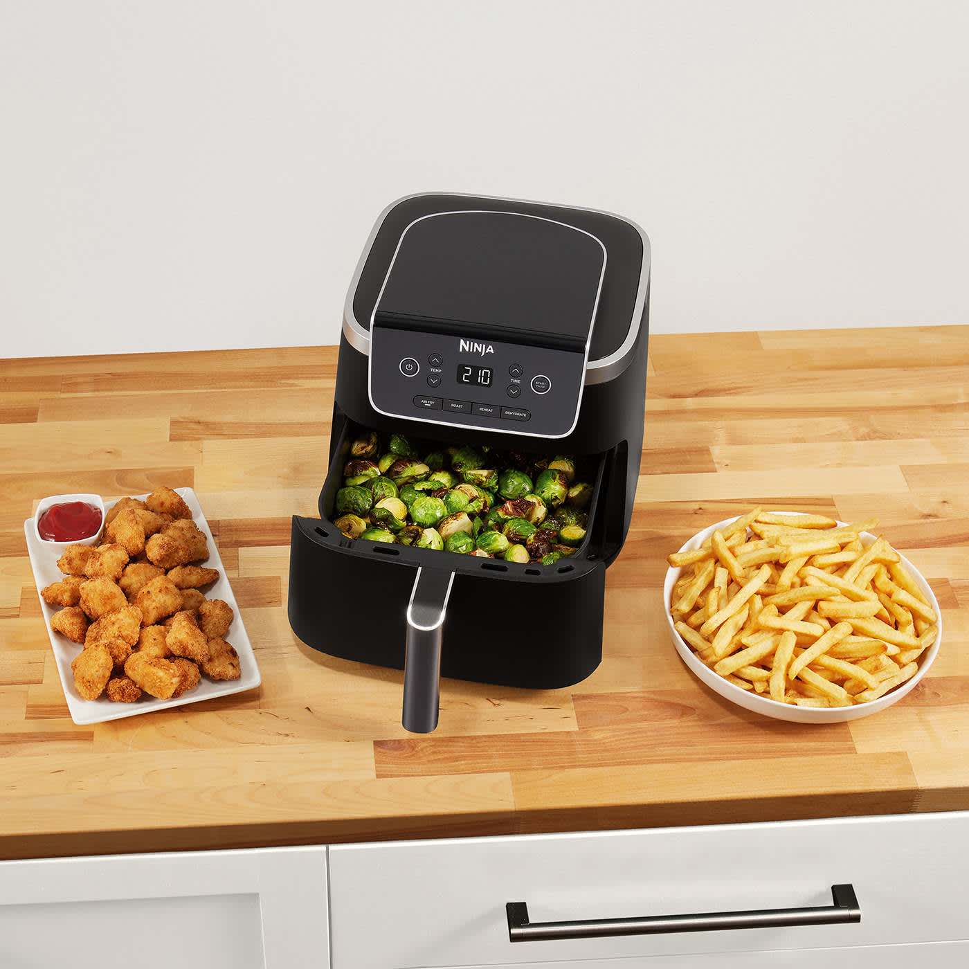 Airfryer avec un tiroir rempli de l&eacute;gumes et entour&eacute; de plusieurs assiettes de nuggets et frites..