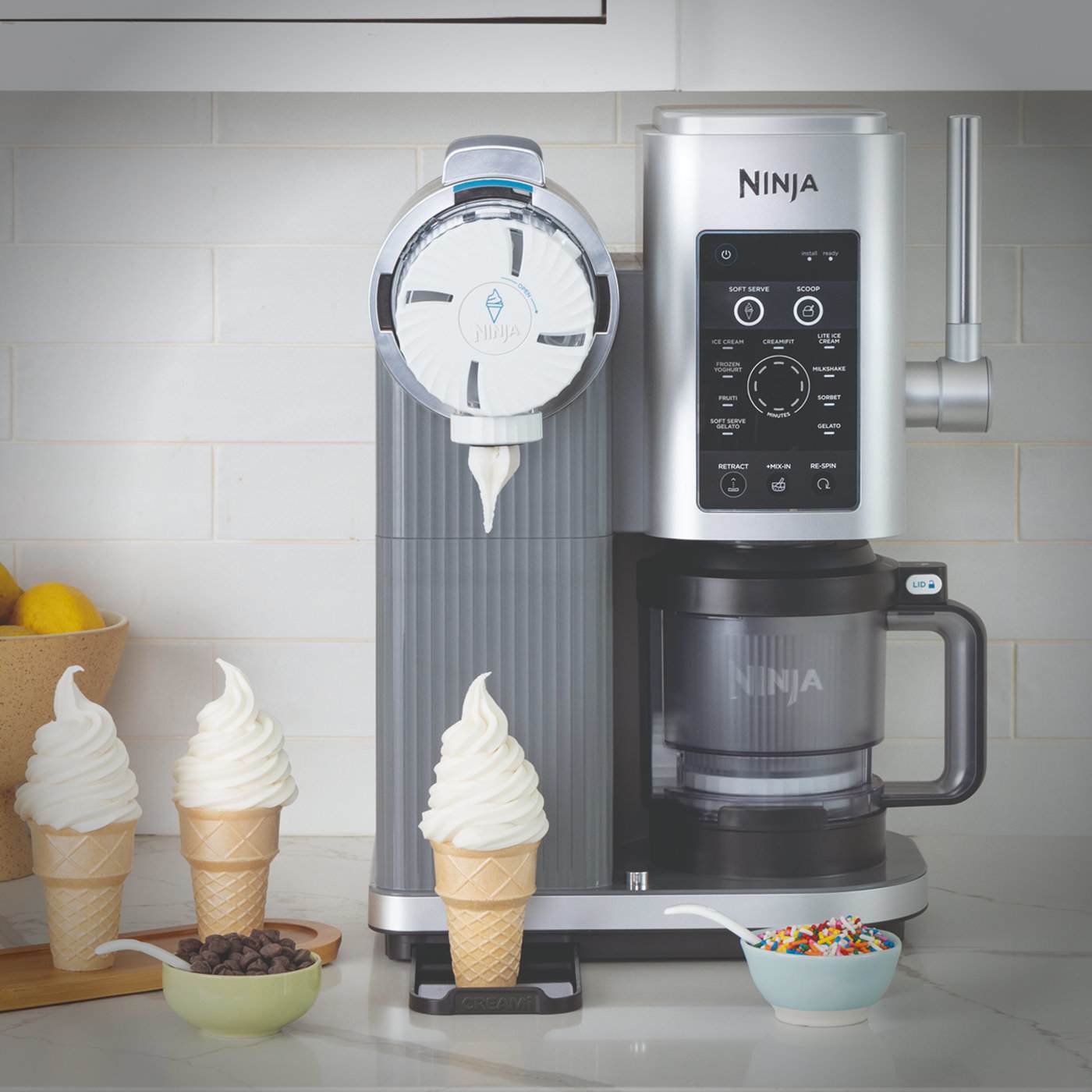 Ninja CREAMi machine dispensing soft serve into an ice cream cone, with cones and toppings arranged beside it on a kitchen worktop.